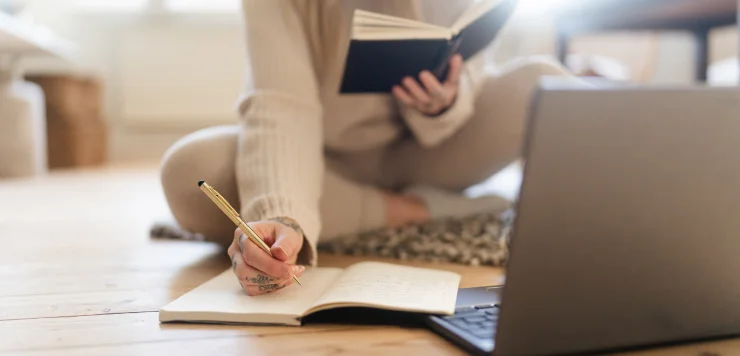 A lady sits on a floor, simultaneously writing and reading in front of her laptop