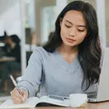 A woman writing on her notepad, with a laptop and phone in front of her