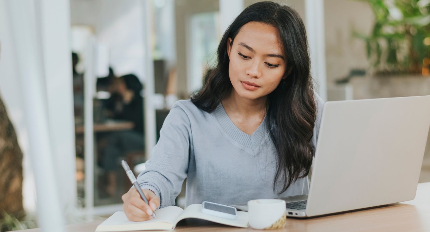 A woman writing on her notepad, with a laptop and phone in front of her